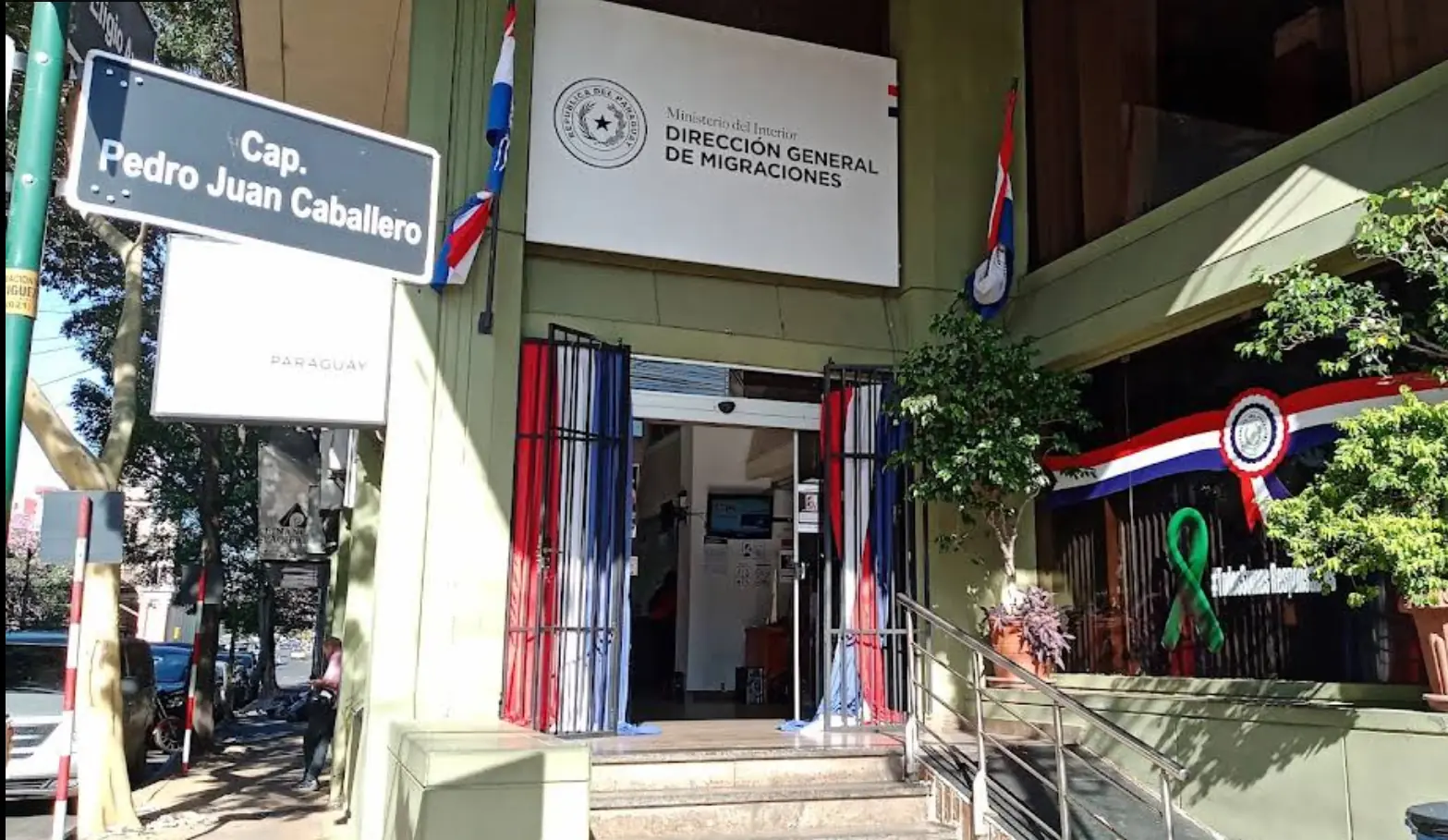 Entrance of the Dirección Nacional de Migraciones in Asunción, with Paraguayan flags and the Cap. Pedro Juan Caballero street sign